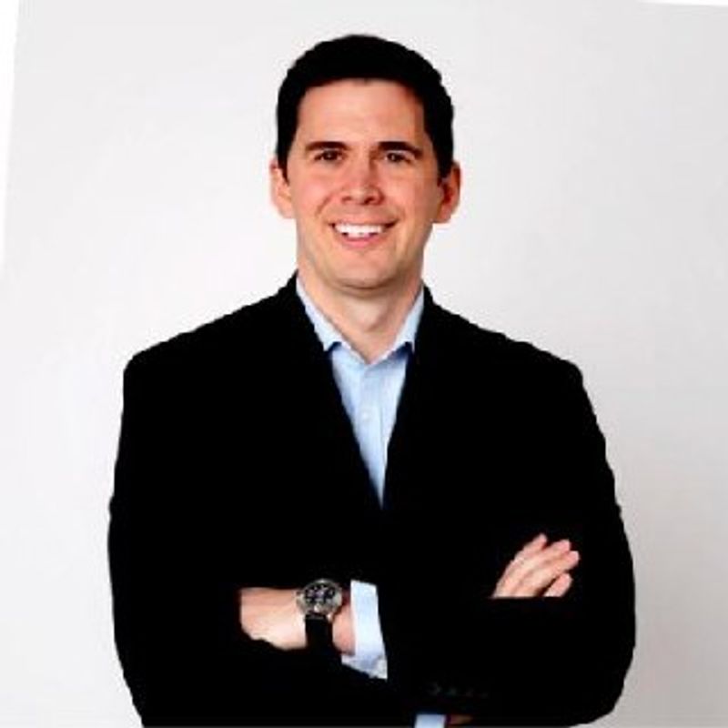 Ethan Hawkes, arms folded, dark jacket over a light blue shirt, white background.